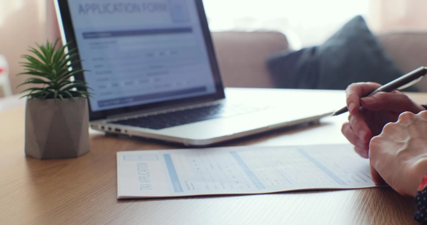 Hands completing a registration form beside a laptop and business documents