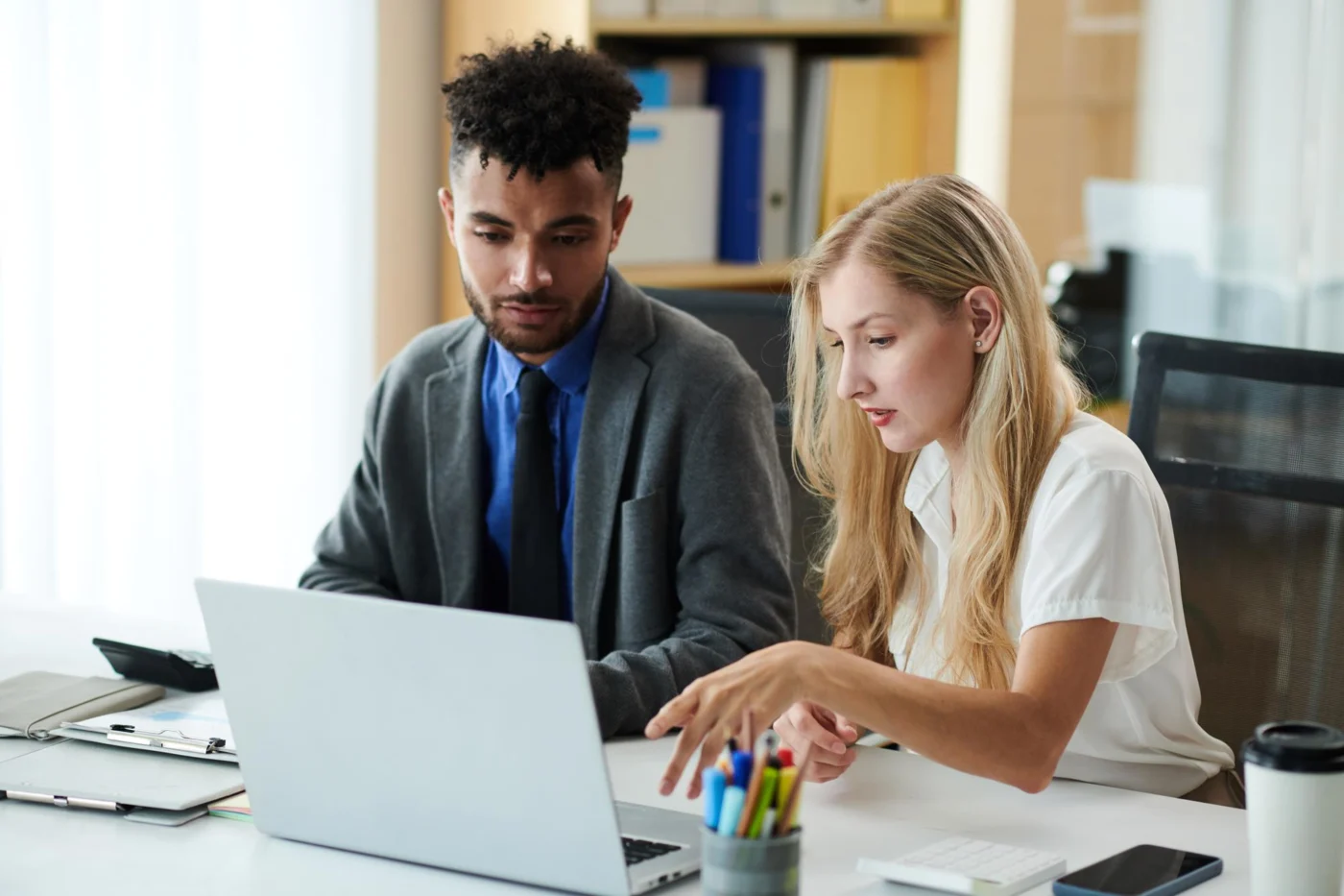 Two colleagues compare figures on a laptop at a bright desk