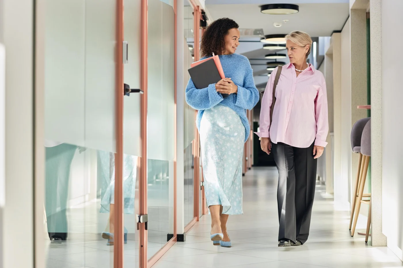 Two colleagues, one senior, chat over a folder in an office corridor
