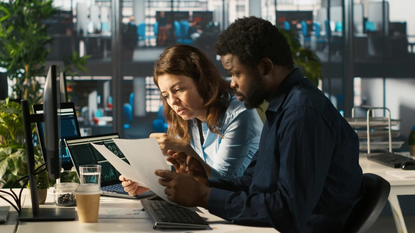 A manager and a colleague check paperwork together at an office desk