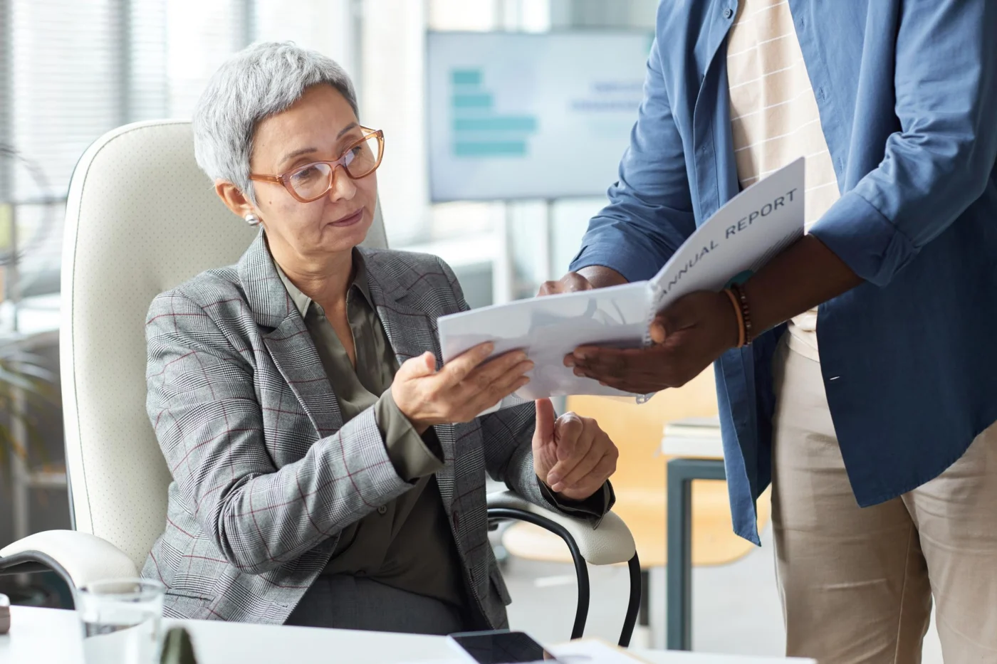 A colleague hands a senior client an annual report to review at her desk